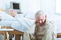 Upset senior man sitting next to elderly woman lying in the ned regarding the seniors' home class actin lawsuit claiming residents died and contracted the illness after operators of CISSS de Laval forced nurses to continue to work at the CHSLD Saint-Dorothée facility even after showing symptoms of the virus.