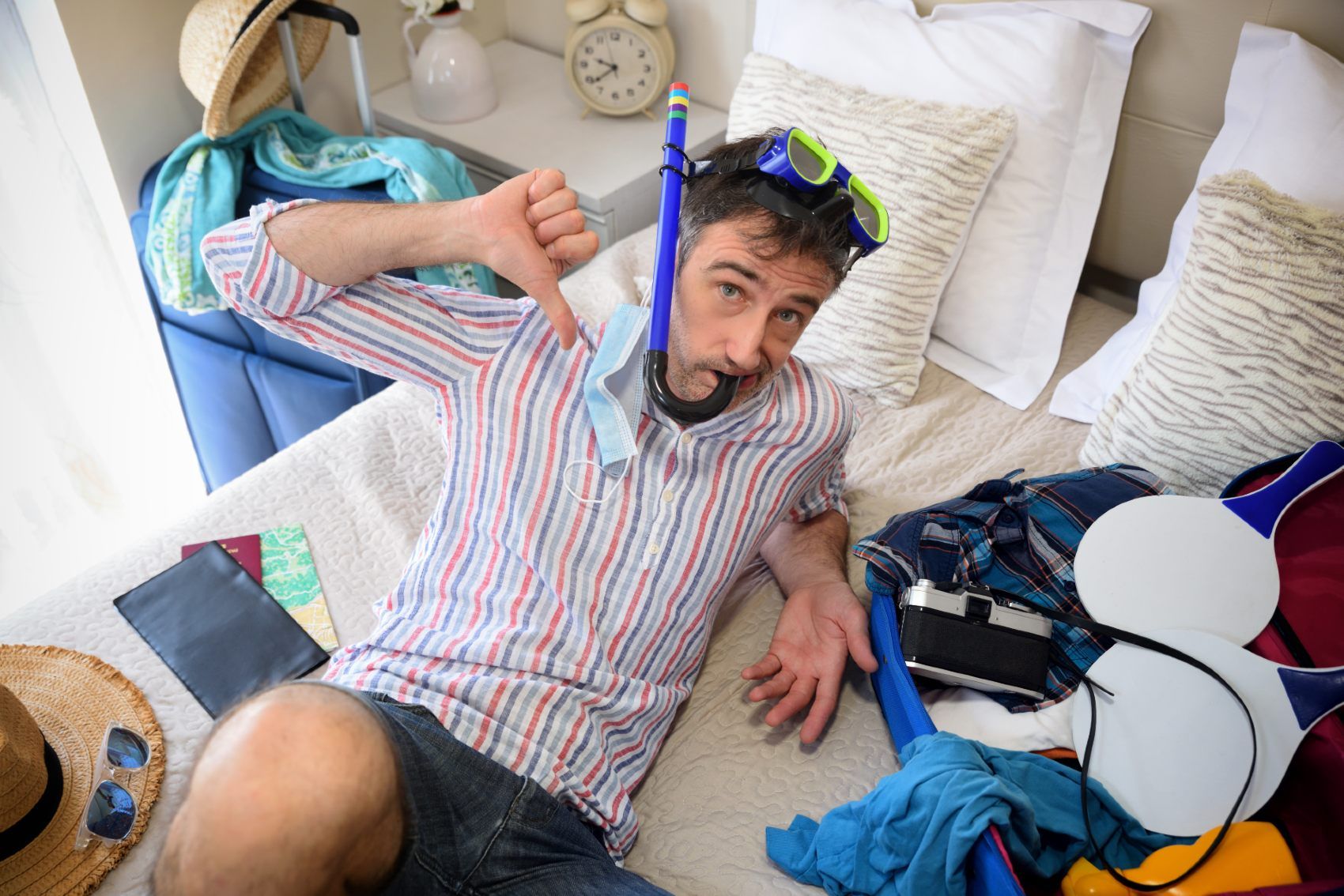 Guy gives a thumbs down while sitting back on his bed while wearing a snorkel, surrounded by open suitcase full of vacation items - td bank