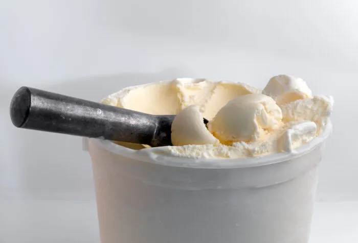 Scoops of vanilla ice cream on top of a tub filled with ice cream next to a metal scoop.