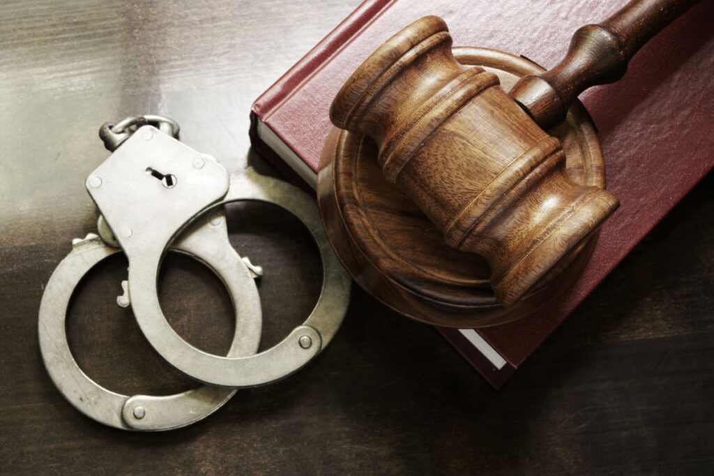 Close up of Judge gavel and handcuffs with red legal book on wooden table.