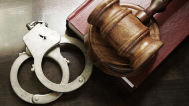 Close up of Judge gavel and handcuffs with red legal book on wooden table.