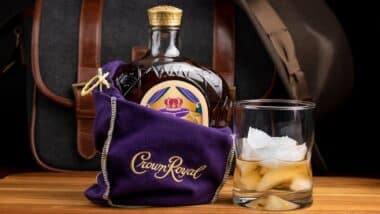 A bottle of Seagram's Crown Royal Canadian rye whiskey in the iconic purple gift bag with a shot in a glass of full of ice cubes.